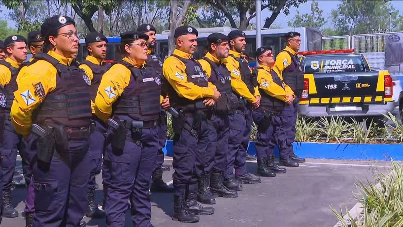 Grupo de guardas municipais uniformizados, com coletes e armas, perfilados ao lado de viatura Força Municipal