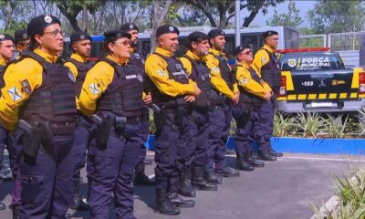 Grupo de guardas municipais uniformizados, com coletes e armas, perfilados ao lado de viatura Força Municipal