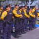 Grupo de guardas municipais uniformizados, com coletes e armas, perfilados ao lado de viatura Força Municipal