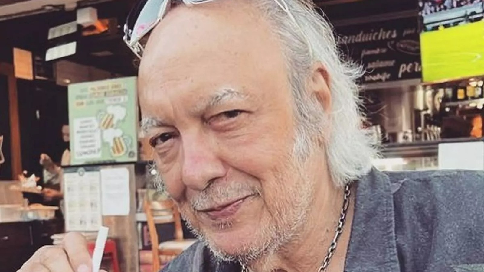Homem de cabelos grisalhos e barba, usando óculos na cabeça e colar de corrente, sorri levemente