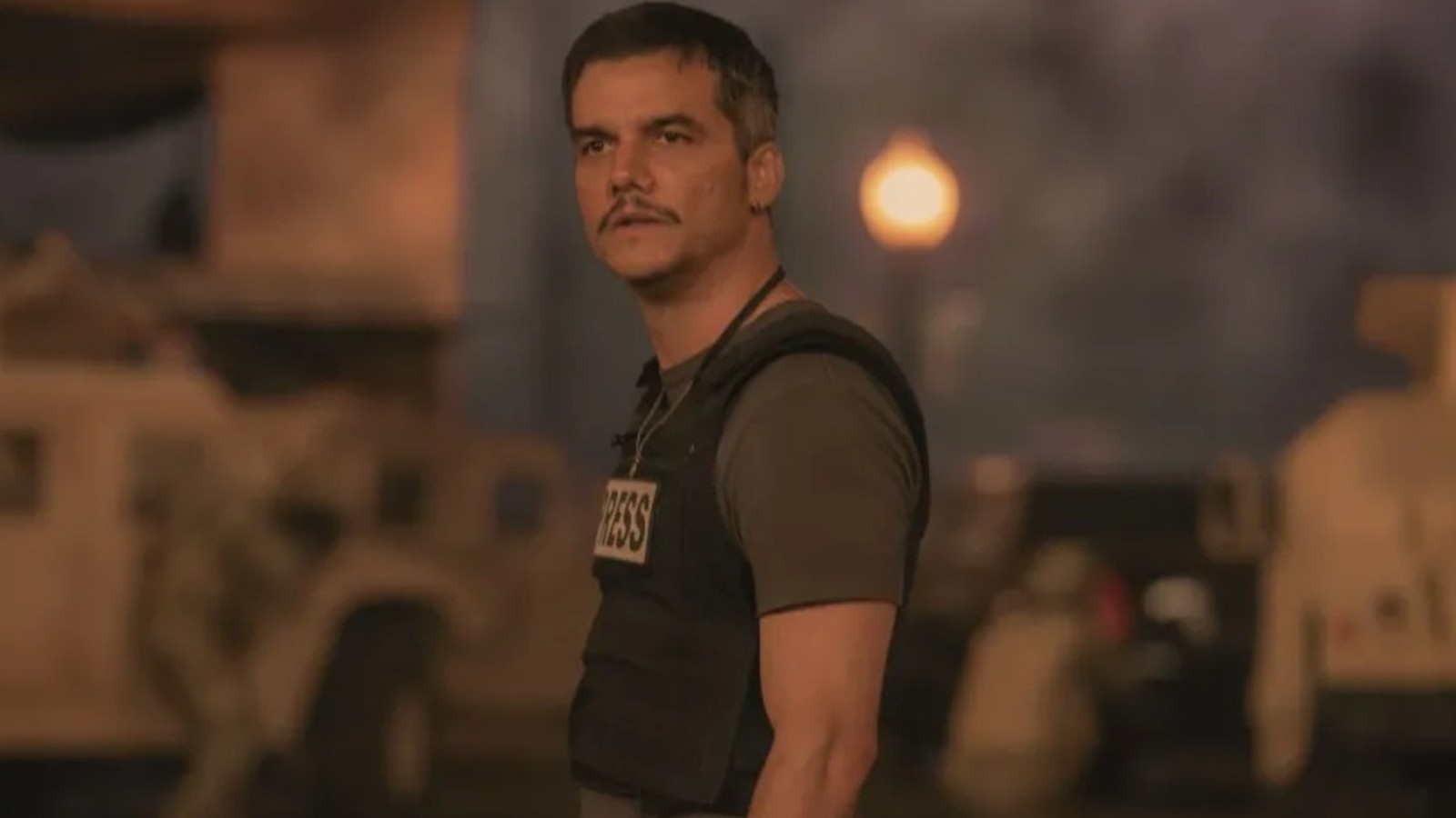 Wagner Moura com bigode e colete de imprensa 'PRESS' em ambiente de combate