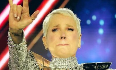 Xuxa Meneghel emocionada em palco, fazendo sinal de 'rock on' com a mão, usando roupa prateada