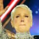 Xuxa Meneghel emocionada em palco, fazendo sinal de 'rock on' com a mão, usando roupa prateada
