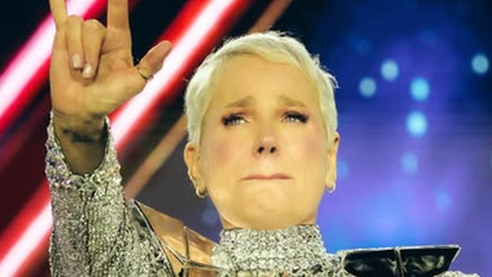 Xuxa Meneghel emocionada em palco, fazendo sinal de 'rock on' com a mão, usando roupa prateada
