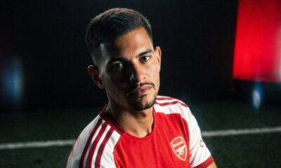 Bruno Guimarães com camisa do Arsenal, olhando para câmera, em fundo escuro com detalhes vermelhos