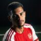 Bruno Guimarães com camisa do Arsenal, olhando para câmera, em fundo escuro com detalhes vermelhos
