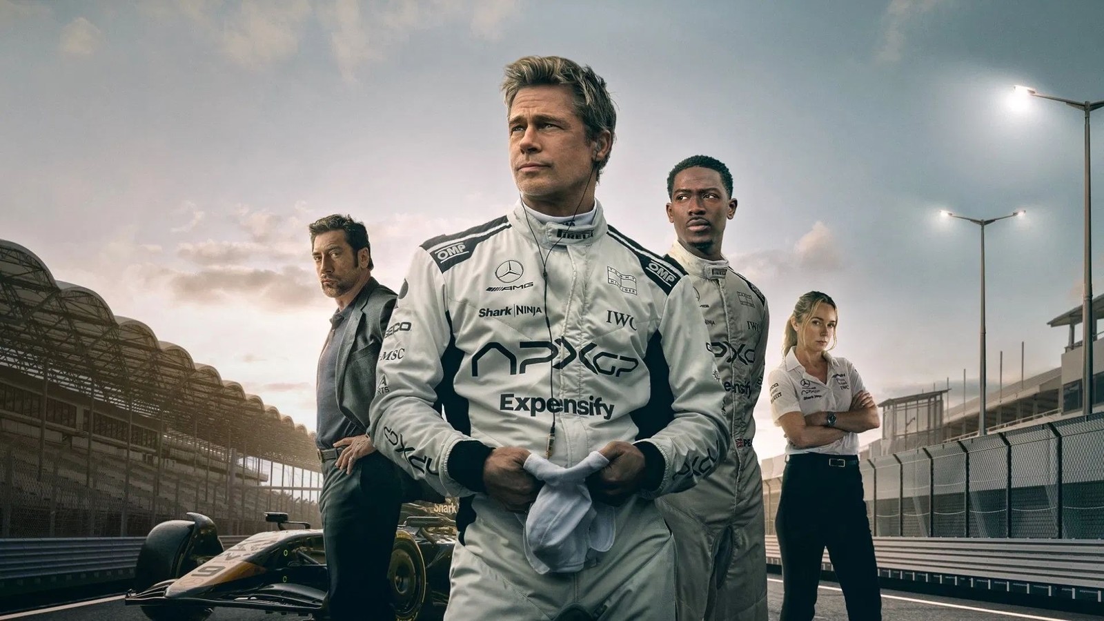 Atores Brad Pitt e Damson Idris em macacões de piloto APX GP na pista, com carro de F1 e equipe