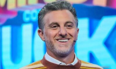 Luciano Huck sorrindo, com cabelo grisalho, barba e suéter listrado marrom e amarelo