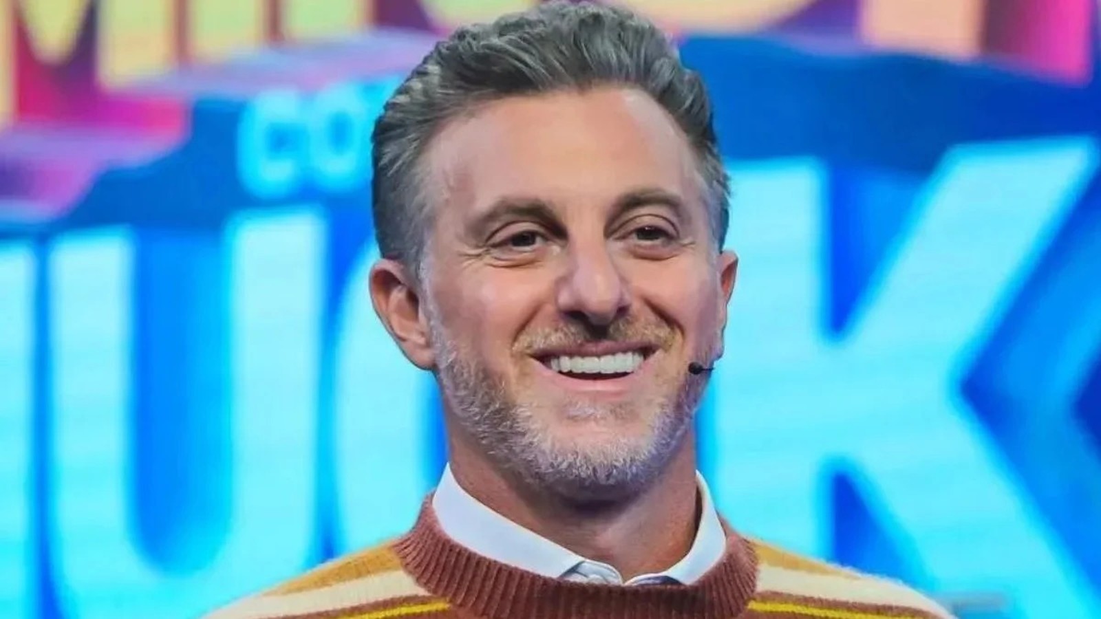Luciano Huck sorrindo, com cabelo grisalho, barba e suéter listrado marrom e amarelo
