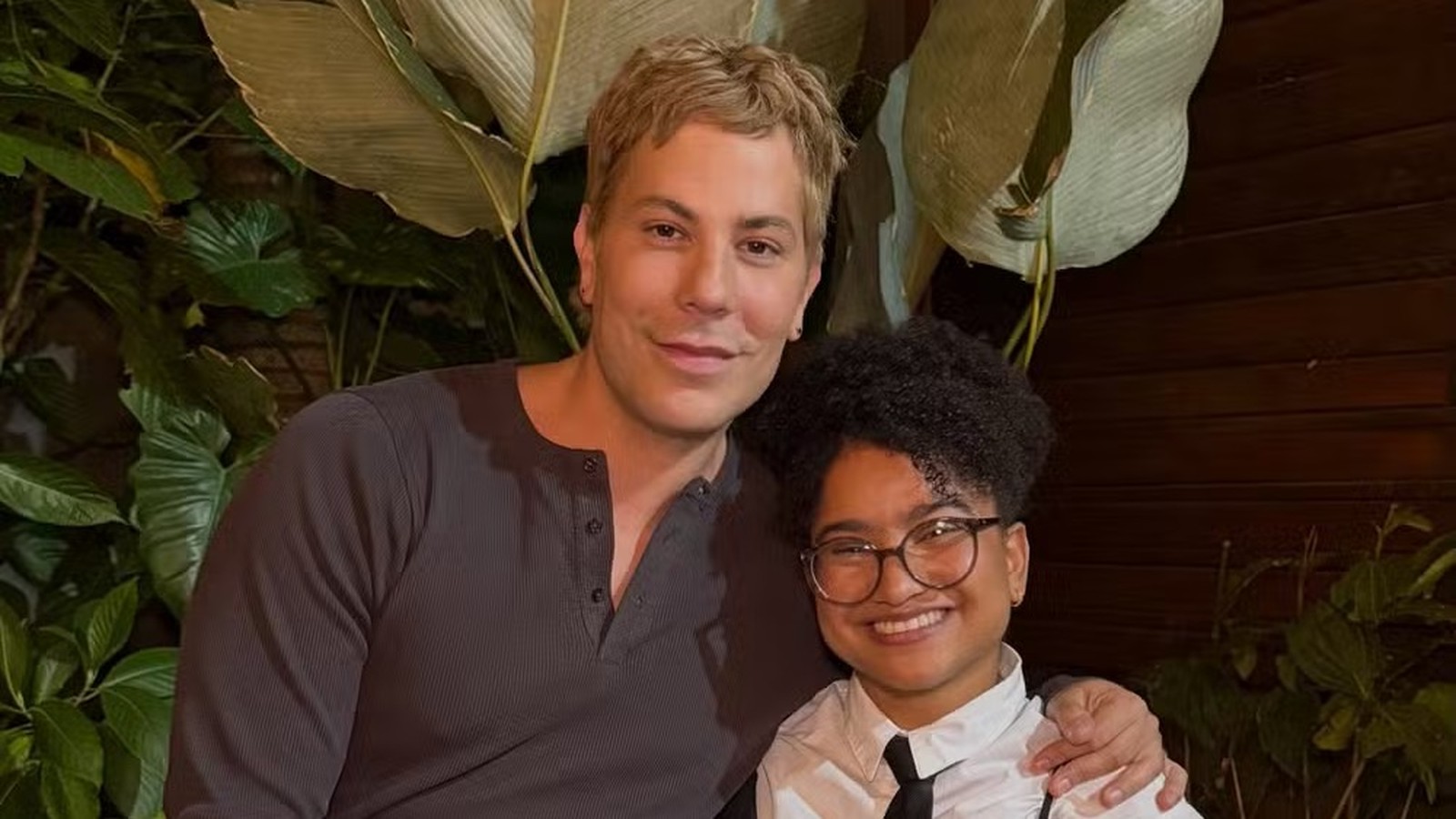Christian Chávez e Tia Milena, sorrindo, em frente a folhagem e parede de madeira