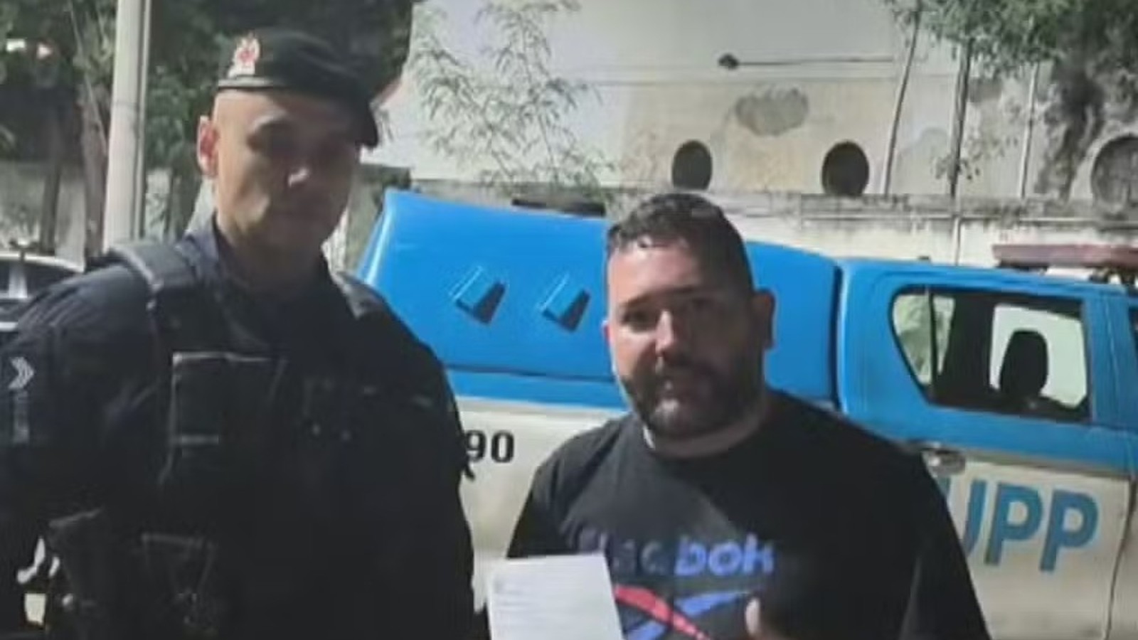 Policial fardado e homem com camiseta Reebok posam à frente de viatura UPP