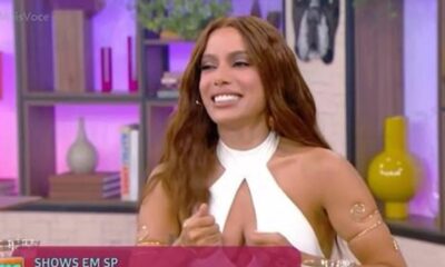 Anitta sorrindo em estúdio de TV, cabelo castanho, top branco e bracelete dourado