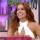 Anitta sorrindo em estúdio de TV, cabelo castanho, top branco e bracelete dourado