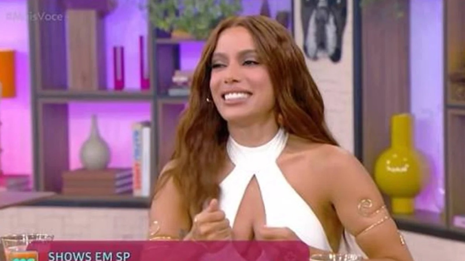 Anitta sorrindo em estúdio de TV, cabelo castanho, top branco e bracelete dourado