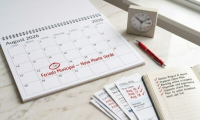 Calendário de agosto de 2026 com o dia 19 destacado como feriado municipal em Nova Monte Verde, ao lado de contas e documentos organizados.