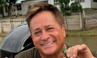 Rosto de Leonardo sorrindo, segurando garrafa, com barco, água e casas ao fundo