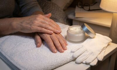 Mãos maduras hidratadas ao lado de creme espesso e luvas brancas de algodão sobre uma toalha.