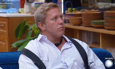 Sérgio Hondjakoff sério, camisa branca, sentado em sofá azul. Fundo cozinha. #RoçaAFazenda, AO VIVO