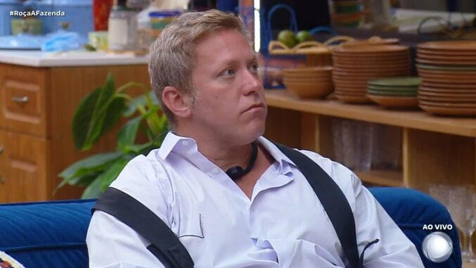 Sérgio Hondjakoff sério, camisa branca, sentado em sofá azul. Fundo cozinha. #RoçaAFazenda, AO VIVO