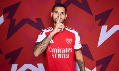 Homem de camisa vermelha do Arsenal com dedo na boca em fundo geométrico vermelho