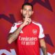 Homem de camisa vermelha do Arsenal com dedo na boca em fundo geométrico vermelho