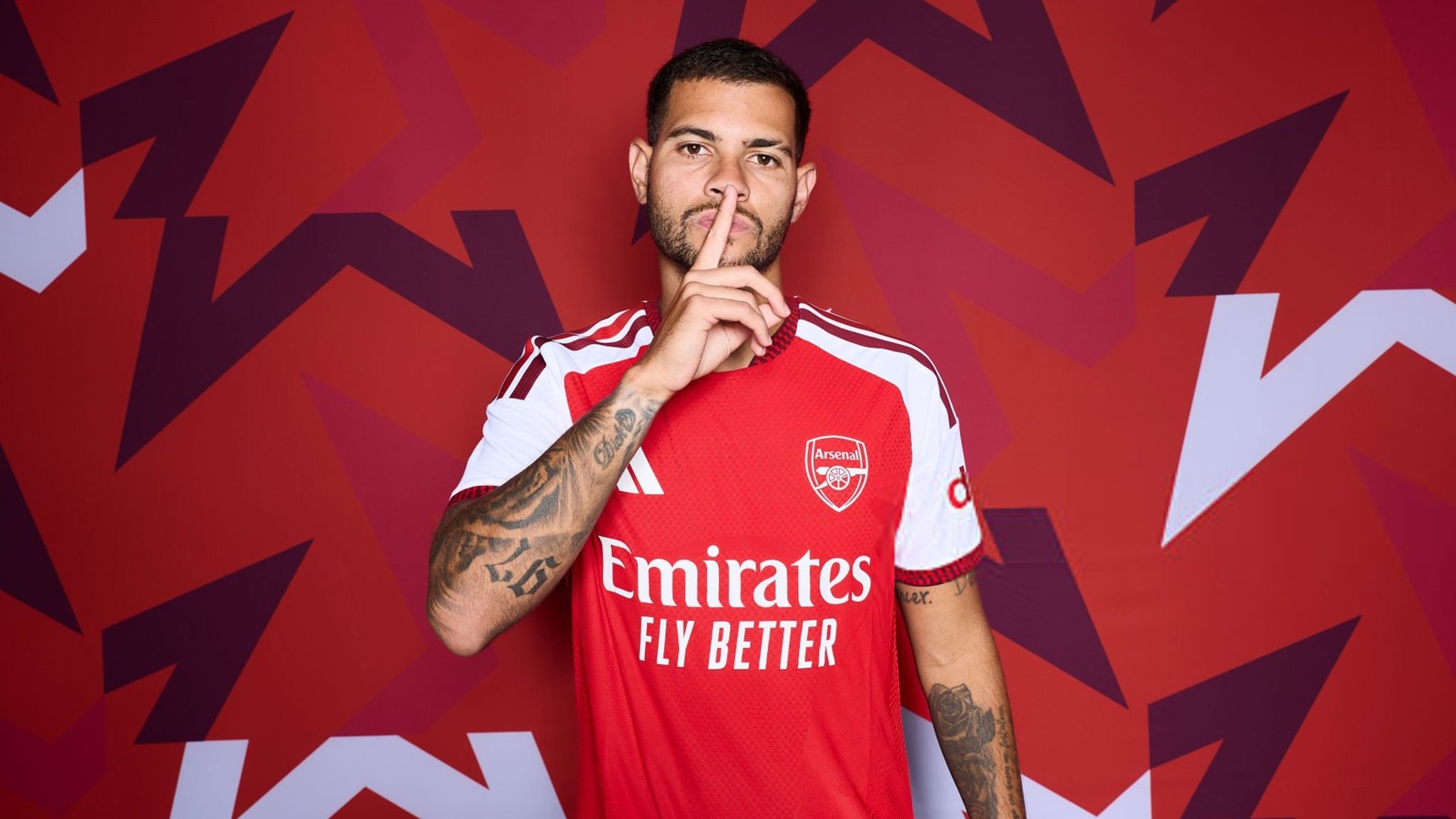 Homem de camisa vermelha do Arsenal com dedo na boca em fundo geométrico vermelho