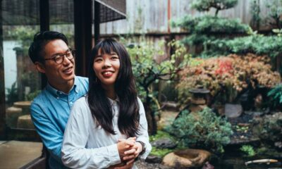Homem e mulher asiáticos sorrindo em abraço, em frente a um jardim japonês com lanterna de pedra