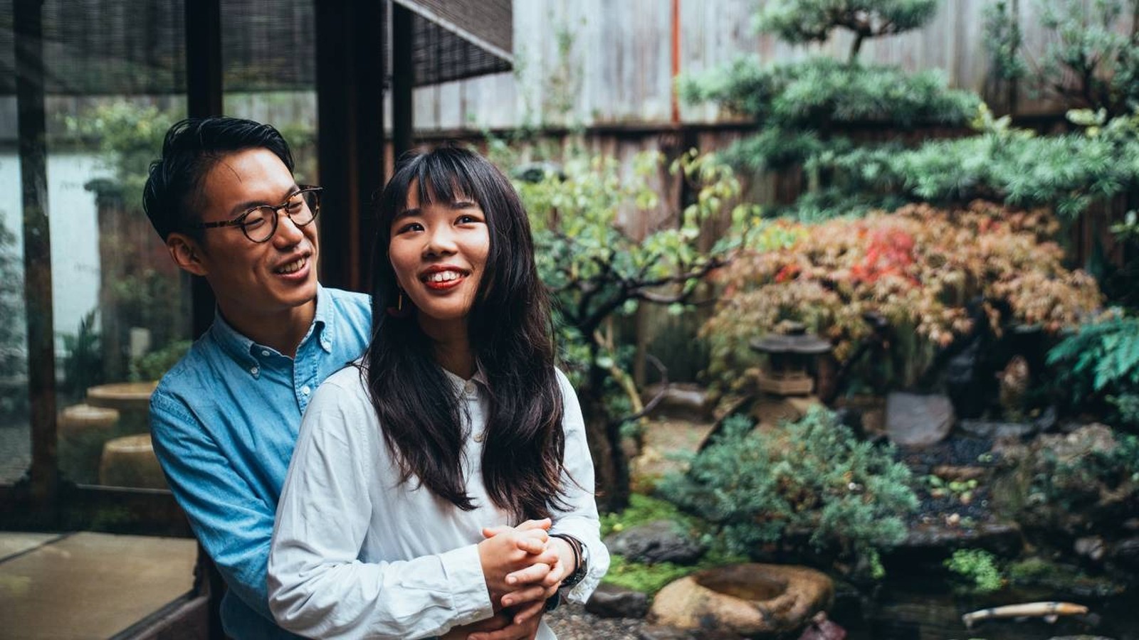 Homem e mulher asiáticos sorrindo em abraço, em frente a um jardim japonês com lanterna de pedra