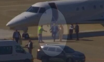 Pessoas descendo de jatinho branco em aeroporto, com van e SUV por perto
