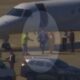 Pessoas descendo de jatinho branco em aeroporto, com van e SUV por perto