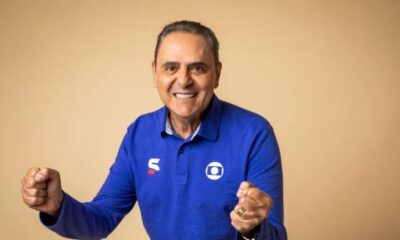 Homem sorridente de camisa azul com logo Globo, punhos cerrados em celebração, fundo bege