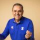 Homem sorridente de camisa azul com logo Globo, punhos cerrados em celebração, fundo bege
