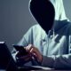 Uma pessoa encapuzada usa um smartphone e um laptop em ambiente escuro