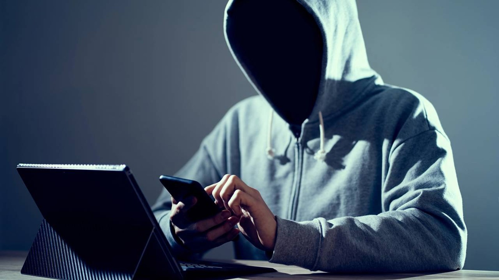 Uma pessoa encapuzada usa um smartphone e um laptop em ambiente escuro