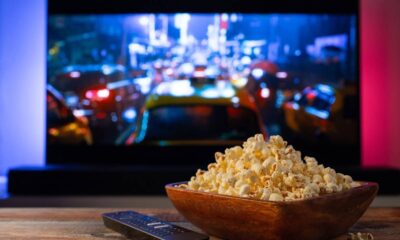 Tigela de pipoca e controle remoto sobre mesa de madeira, TV exibe cena de rua noturna