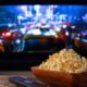 Tigela de pipoca e controle remoto sobre mesa de madeira, TV exibe cena de rua noturna