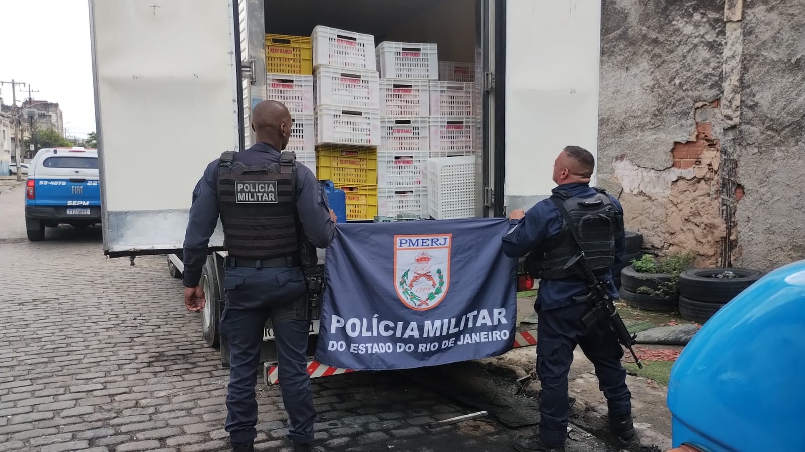Dois policiais seguram bandeira da PMERJ; atrás, caminhão com caixas Kerfrango e viatura policial