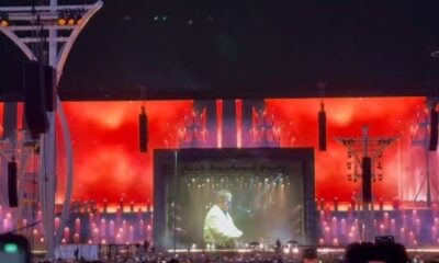 Palco de show com tela exibindo Elton John em jaqueta clara, luzes vermelhas e elementos que remetem a velas