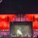 Palco de show com tela exibindo Elton John em jaqueta clara, luzes vermelhas e elementos que remetem a velas