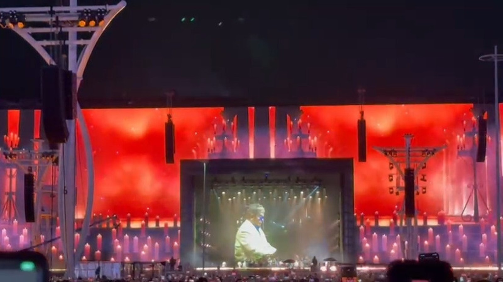 Palco de show com tela exibindo Elton John em jaqueta clara, luzes vermelhas e elementos que remetem a velas