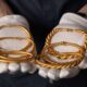 Mãos usando luvas brancas seguram vários braceletes de ouro, alguns lisos e outros torcidos
