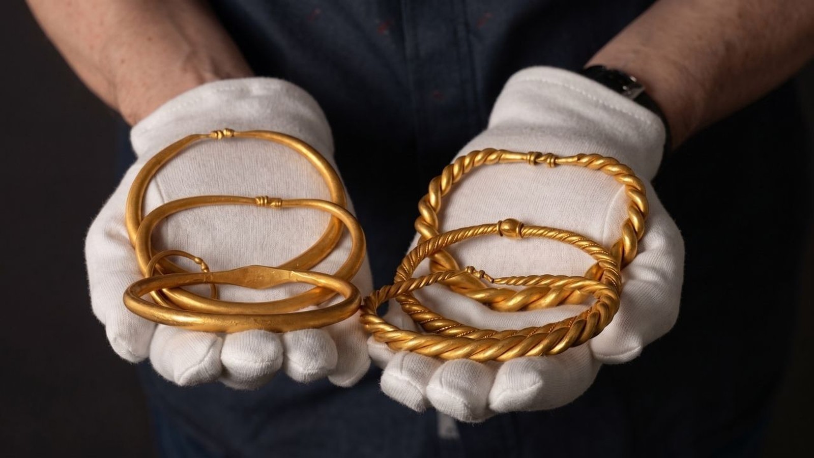 Mãos usando luvas brancas seguram vários braceletes de ouro, alguns lisos e outros torcidos