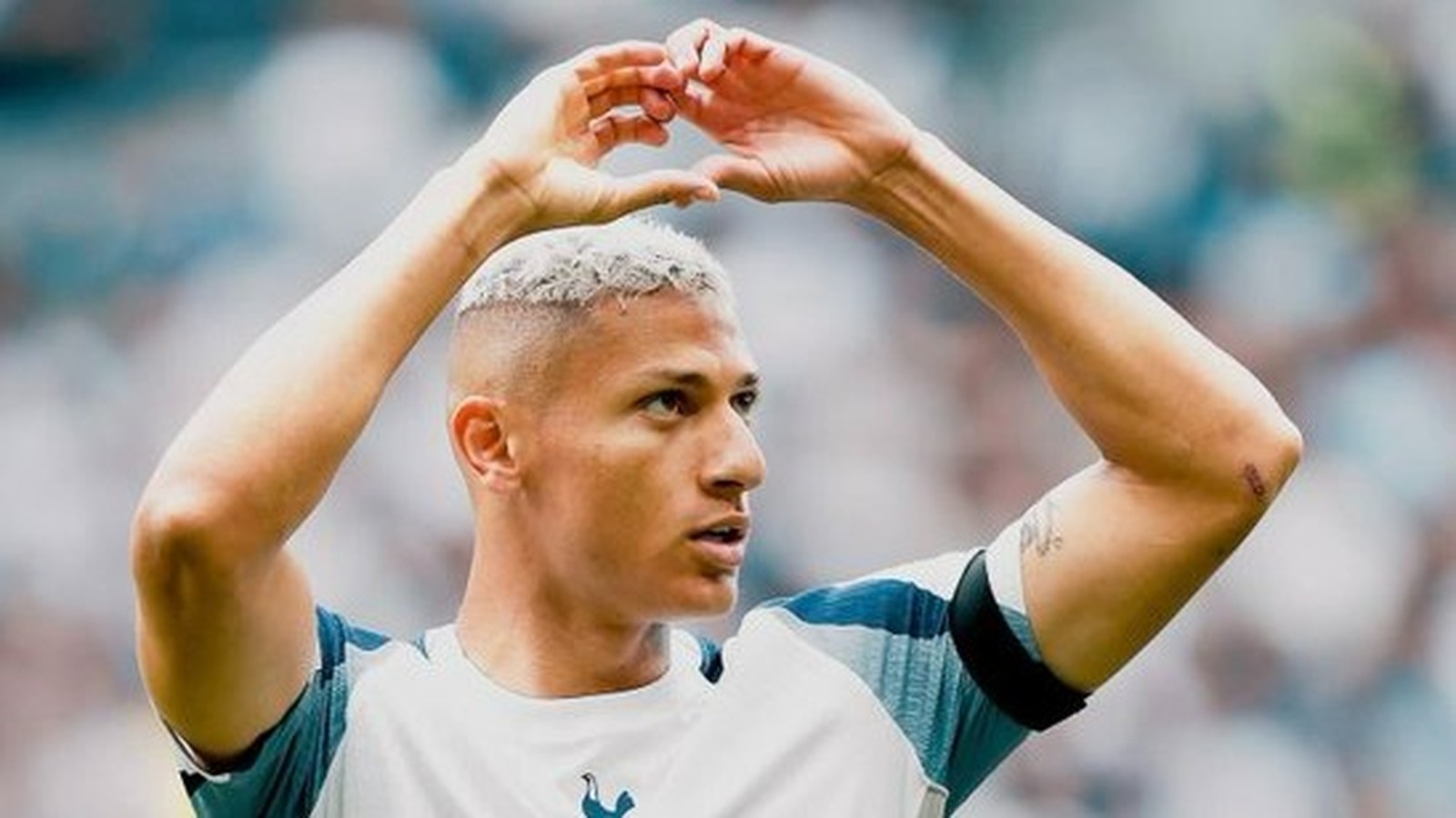 Richarlison com cabelo platinado faz gesto de coração com as mãos em campo de futebol