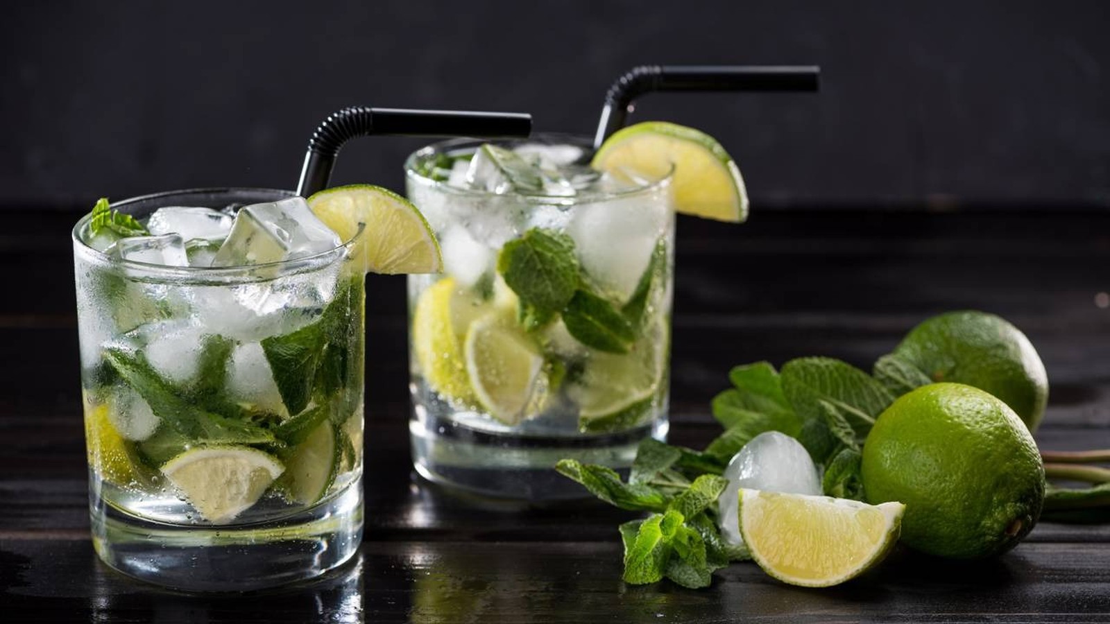 Dois copos de mojito com gelo, hortelã, limão, canudos pretos em mesa escura