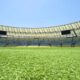 Gramado verde de estádio vazio com assentos azuis e amarelos, telão e céu azul