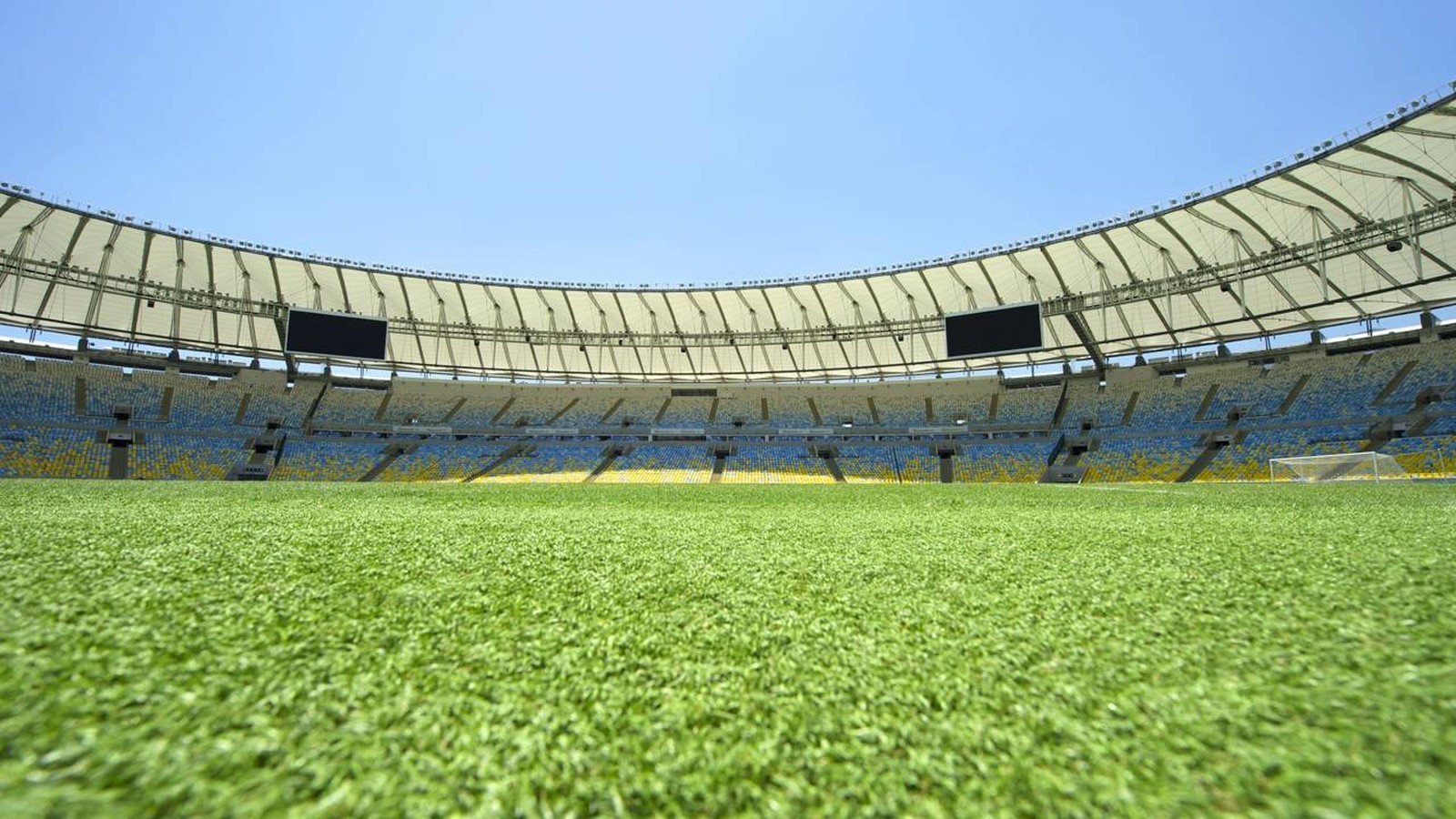 Gramado verde de estádio vazio com assentos azuis e amarelos, telão e céu azul