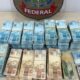 Pilhas de notas de R$100 e R$50 reais sobre mesa com escudo da Polícia Federal
