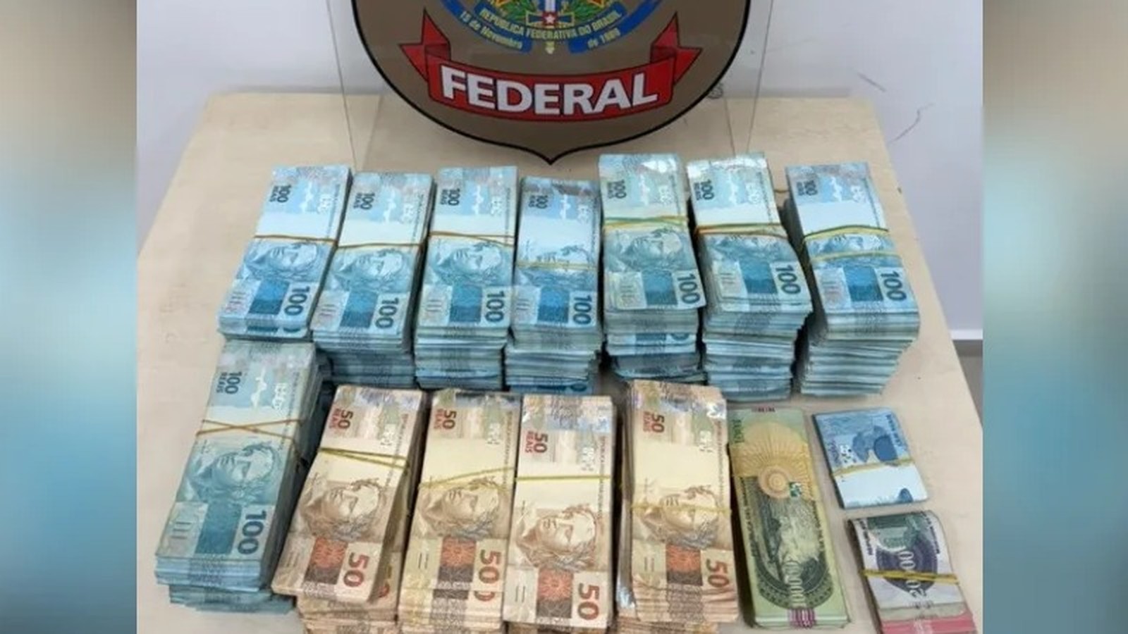Pilhas de notas de R$100 e R$50 reais sobre mesa com escudo da Polícia Federal