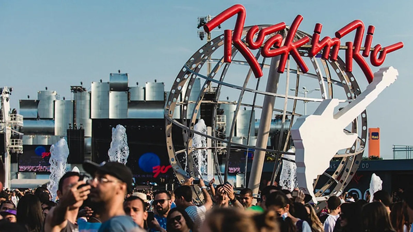 Multidão em festival, com letreiro vermelho 'Rock in Rio', guitarra branca e globo metálico