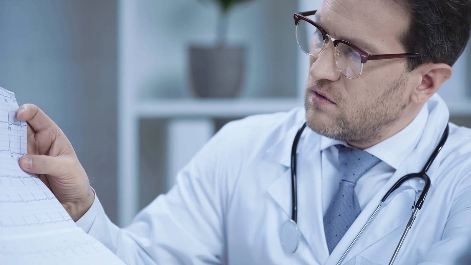 Médico com óculos e estetoscópio examinando atentamente um eletrocardiograma